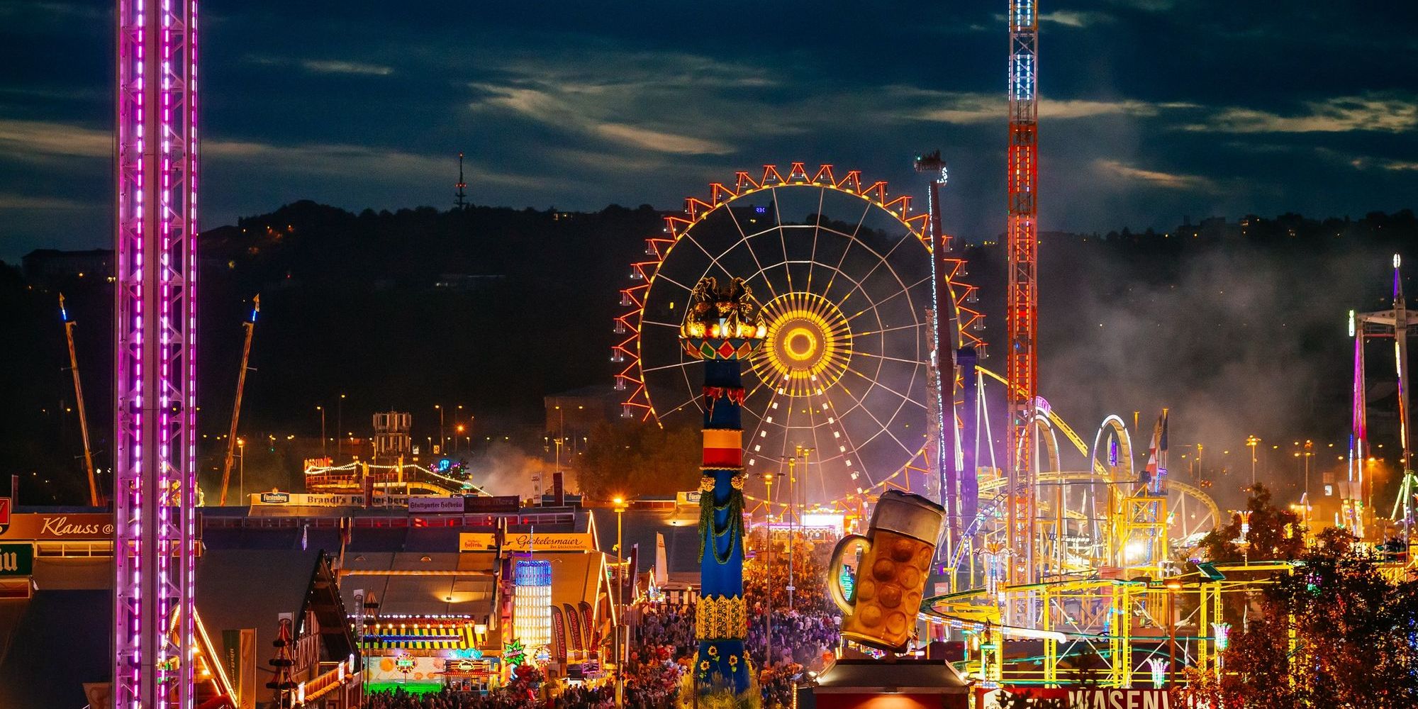 Blick auf das Volksfest aus der Vogelperspektive mit den Bierzelten, dem Freefall-Tower, Infinitiv, dem Riesenrad, einer Achterbahn und vielen Menschen auf den Wegen zwischen den Zelten.