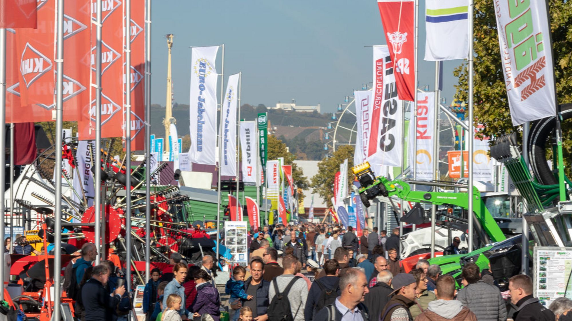 Blick auf einen Weg des Messegeländes mit vielen Menschen, Fahnenstangen und Fahrzeugen