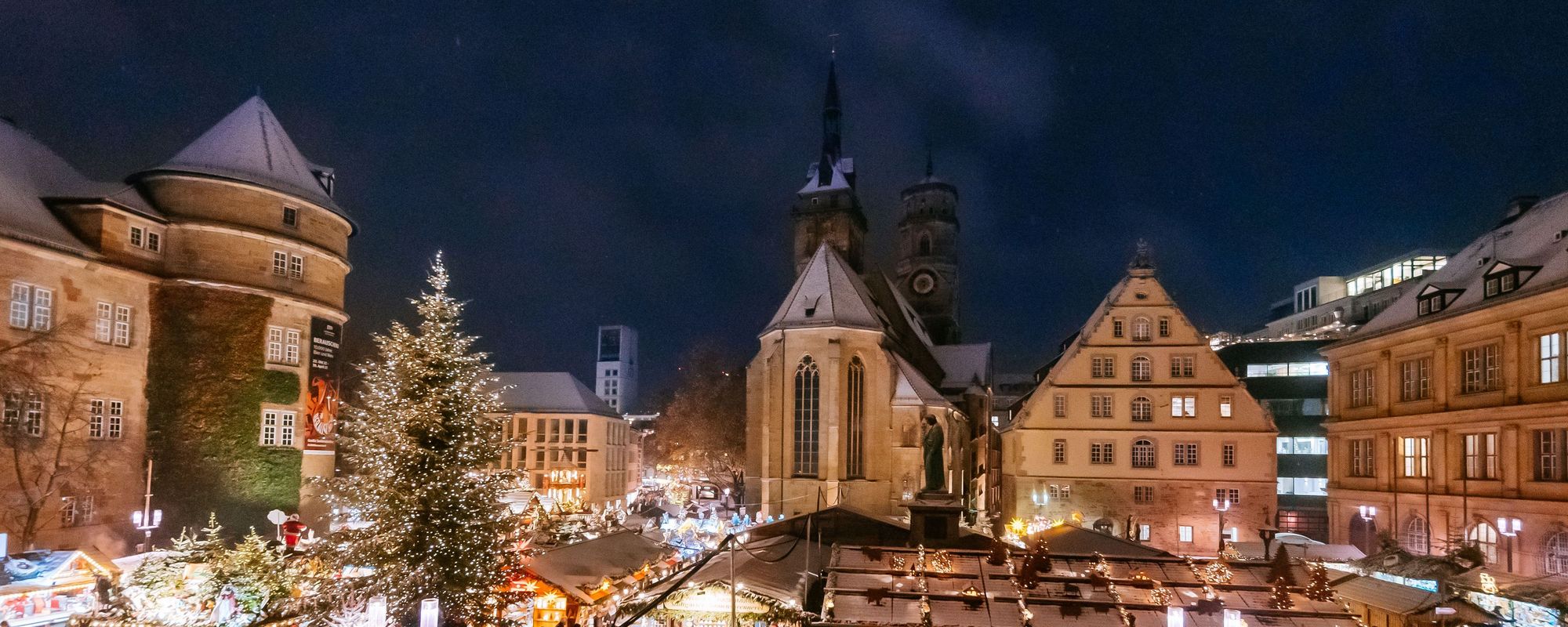 Blick auf den Rathausplatz während des Weihnachtsmarktes. Die Buden haben Schnee auf den Dächern und leuchten in der Dämmerung